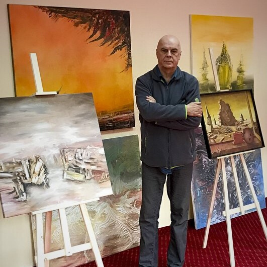 Zygmunt Jasnoch stoi poważnie z założonymi rękami, pośród swoich obrazów na ścianach i sztalugach, tuż przed wystawą.